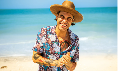 Niko Moon clasps his hands together and stands in front of a sandy beach.