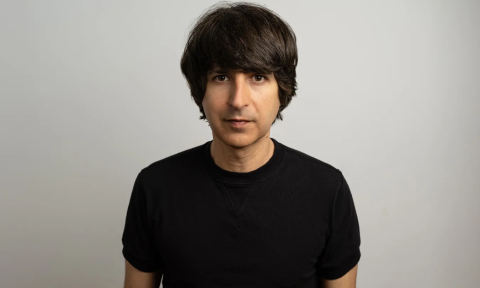 Demetri Martin dons a black shirt and poses in front of a gray backdrop.