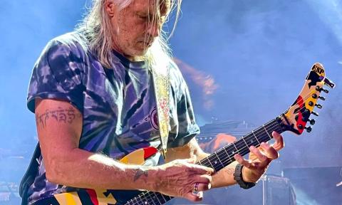 George Lynch clutches his guitar and performs on stage in front of a blue backdrop.