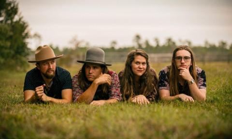 Members of Remedy Tree lie flat on their stomachs, posing playfully atop a lush bed of grass.