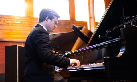 Pianist Aaron Lehrian sitting at a grand piano in a wooden paneled room with large upper windows