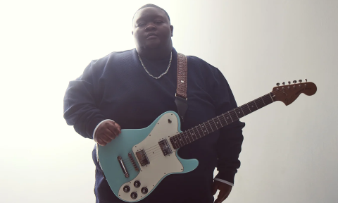 Christone "Kingfish" Ingram clutches his guitar and poses in front of a white backdrop.