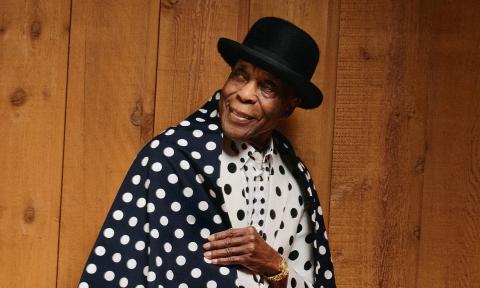 Buddy Guy stands for the camera in a black hat and a polka-dot jacket.