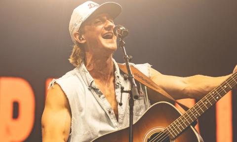 Cooper Alan wears a hat and strums his guitar on stage. 