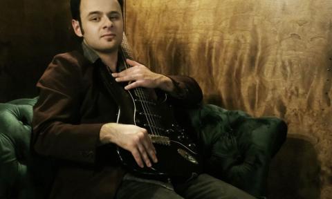 Yates McKendree poses with his guitar in front of a brown backdrop.