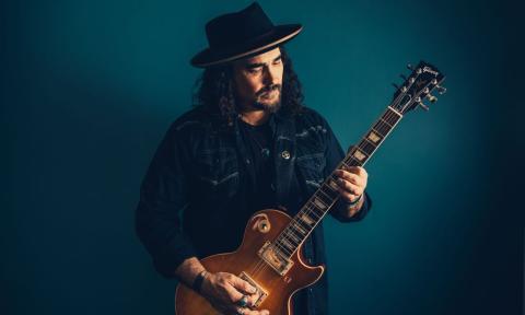 Alastair Greene wears a black hat and strums his guitar in front of a blue backdrop.