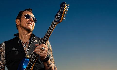 Gary Hoey strums his guitar while wearing black sunglasses.
