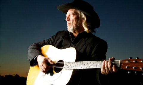 John Anderson dons a black cowboy hat and strums his guitar. 