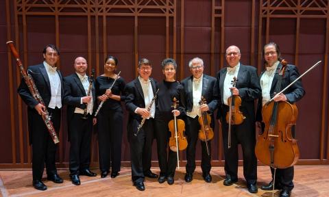 Eight musicians in a winds and strings ensemble, standing in a room with majestic wood paneling