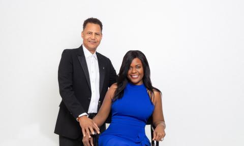 A couple, the man is wearing black and white and standing, the woman is seating and wearing a bright blue dress