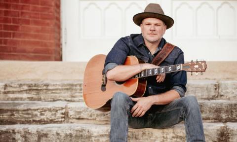 Jason Eady sits on a staircase and poses with his guitar.