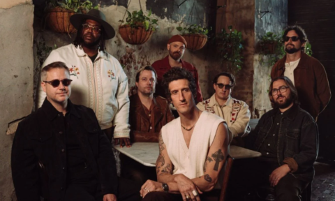 Members of The Revivalists stand together before a textured backdrop accented with hanging pots.