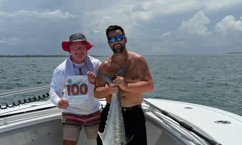 Two men on the foredeck of a fishing holding a kingfish and a sign with their boat's number