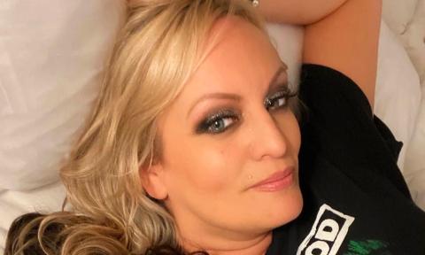 Stormy Daniels is dressed in a black top, reclining on white sheets with a coordinating pillow.