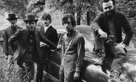 The Band’s members, dressed in dark attire, cluster together beside a vintage car.