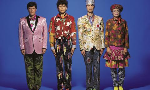 The Talking Heads wear colorful clothing and pose in front of a blue backdrop.