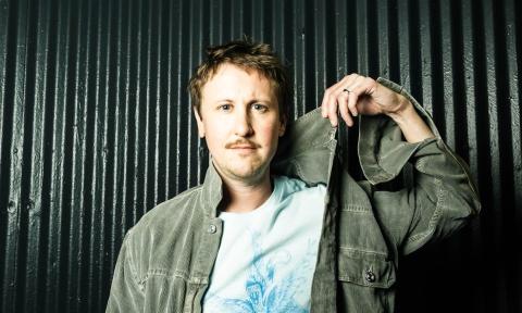 Johnny Pemberton grabs his green shirt and poses in front of a dark green backdrop.