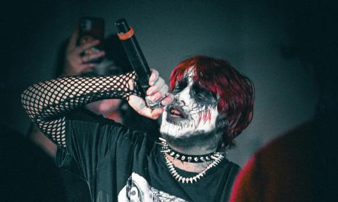 A singer cloaked in gothic attire and striking makeup commands the stage with a haunting live performance.