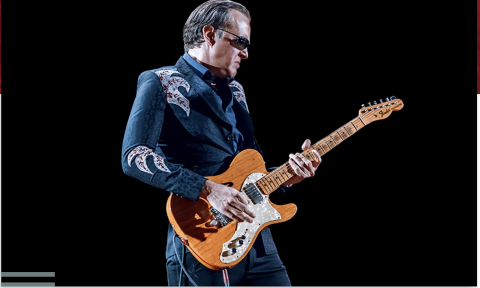 Joe Bonamassa strums his guitar in front of a black backdrop.
