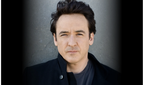 John Cusack wears black clothing and poses in front of a black and grey backdrop.