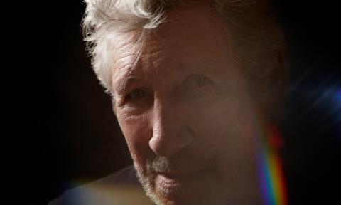 A headshot of Roger Waters as he poses in front of a black backdrop.