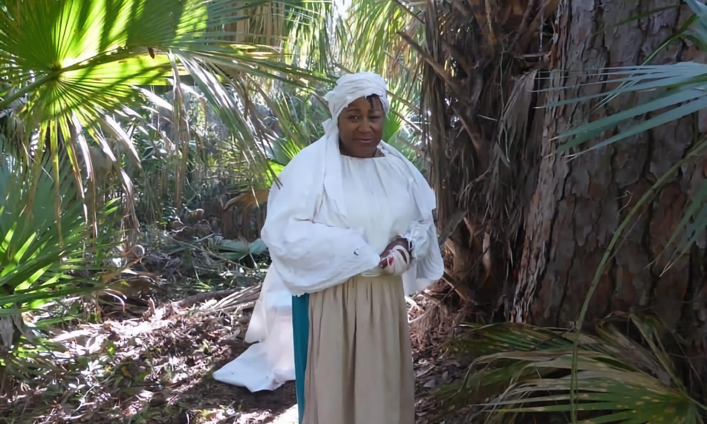 A black-American woman in the dress of the 1700s standing in a forest clearing