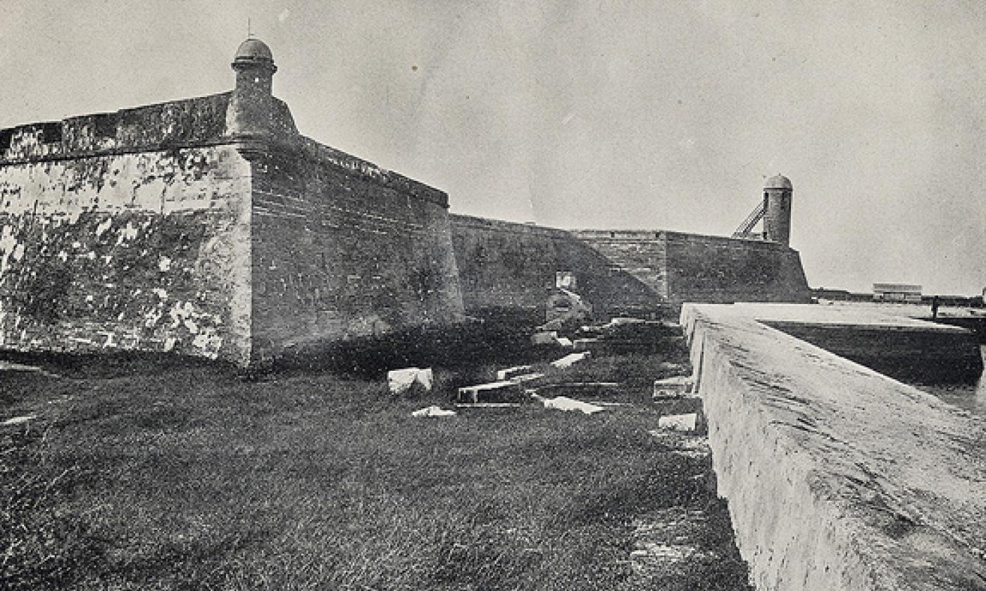 A black and white photograph of the Castillo de San Marcos (then Fort Marion) in St. Augustine Florida. The photo faces north and features the sea wall along Matanzas Bay on the right side of the image. The coquina for stands in a 'moat' full of grass.