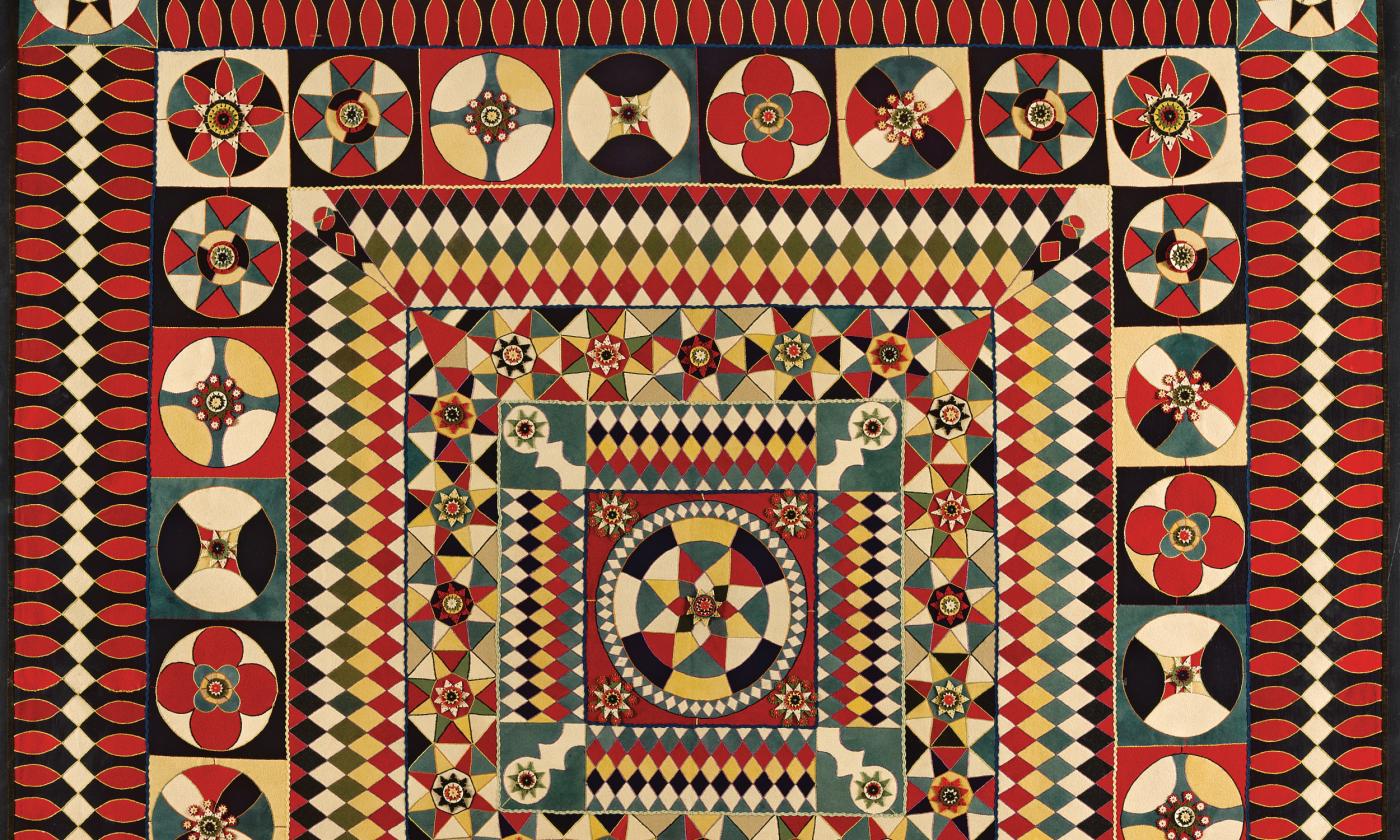 A detail view of an old quilt, predominately red, cream, and blue in color, and an intricate pattern that changes from the edges to the center
