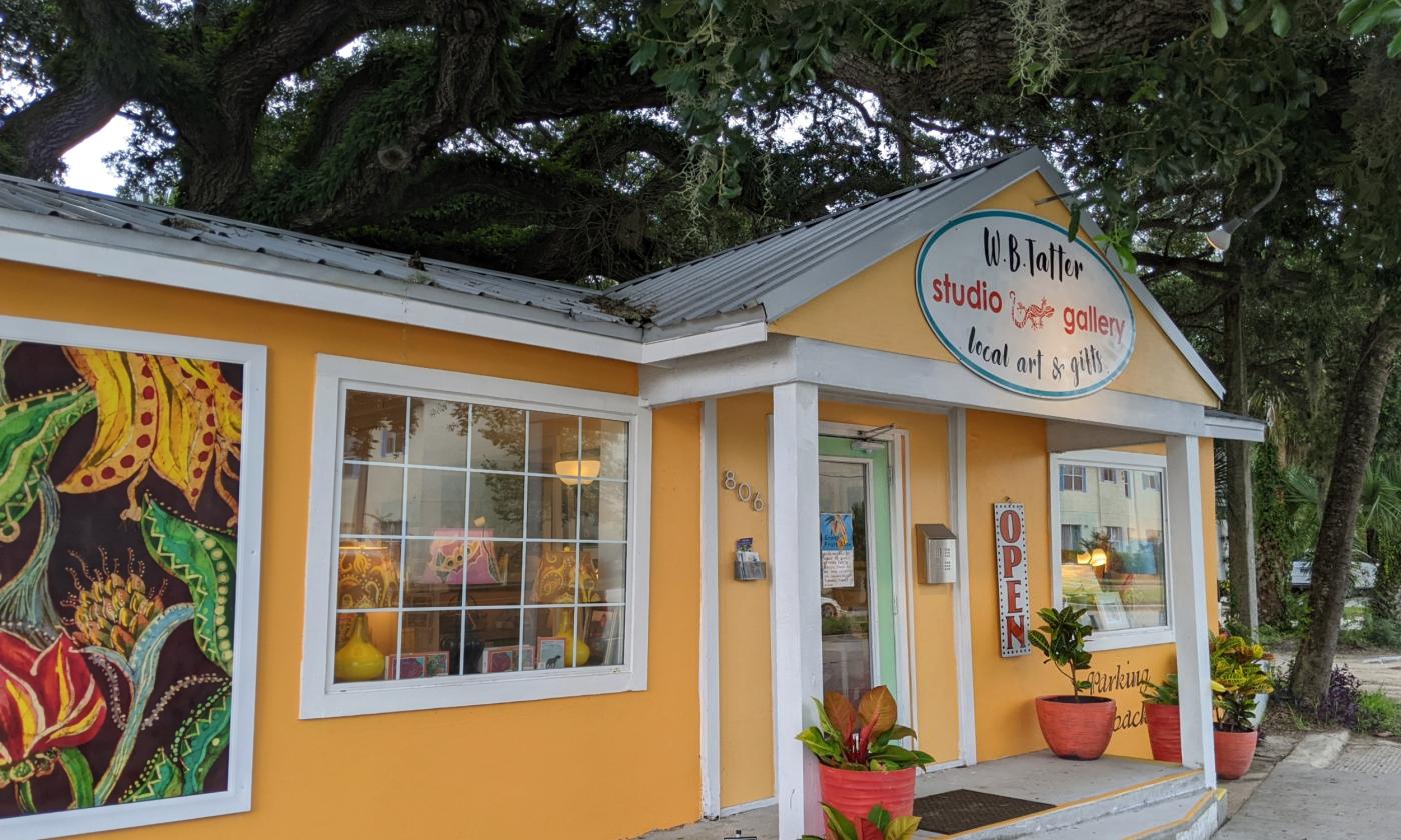 A small building, painted yellow, with plants and art outside, and a sign for a gallery