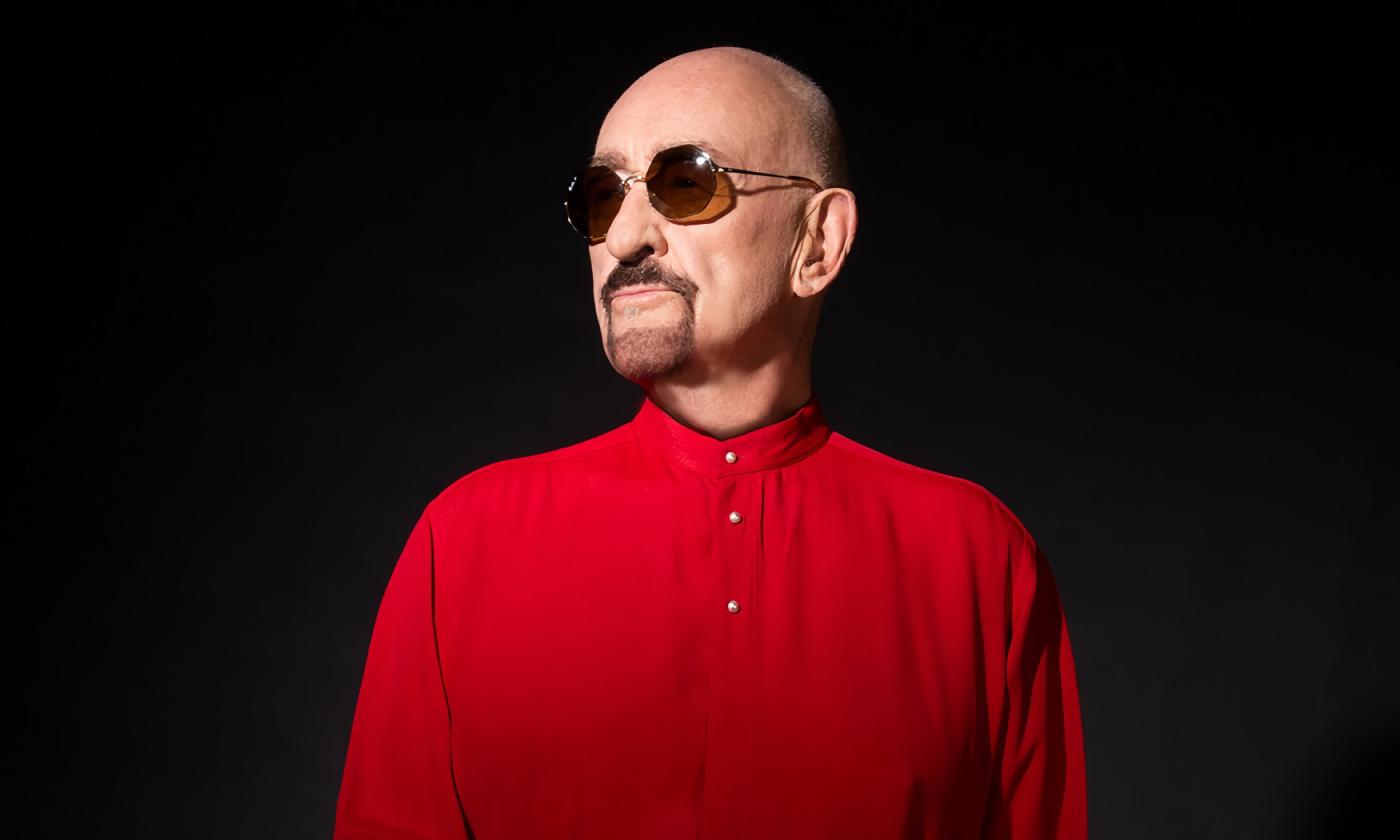 Musician Dave Mason stands in a red shirt and sunglasses, against a black background, looking to his right.