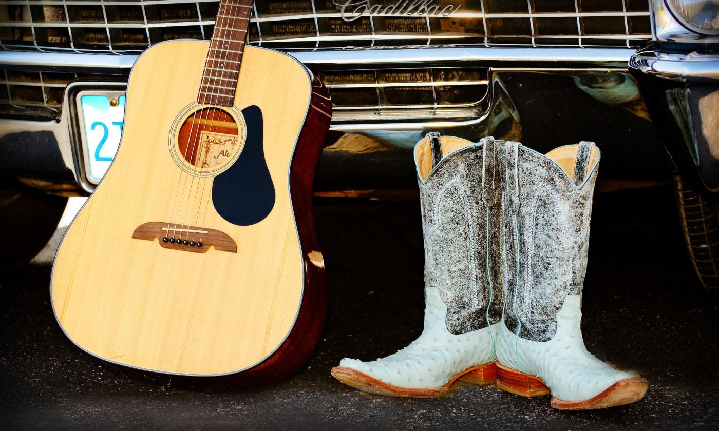 A Pixabay photo with a guitar standing up in front of the grill of an old Cadillac next to a pair of white cowboy boots