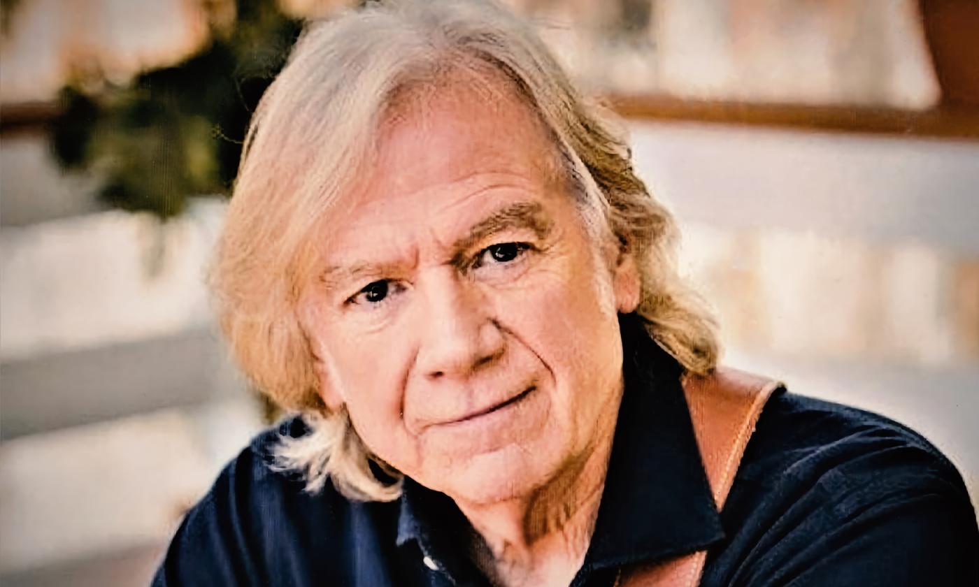 Justin Hayward, in black shirt with guitar strap over his shoulder, against a white building