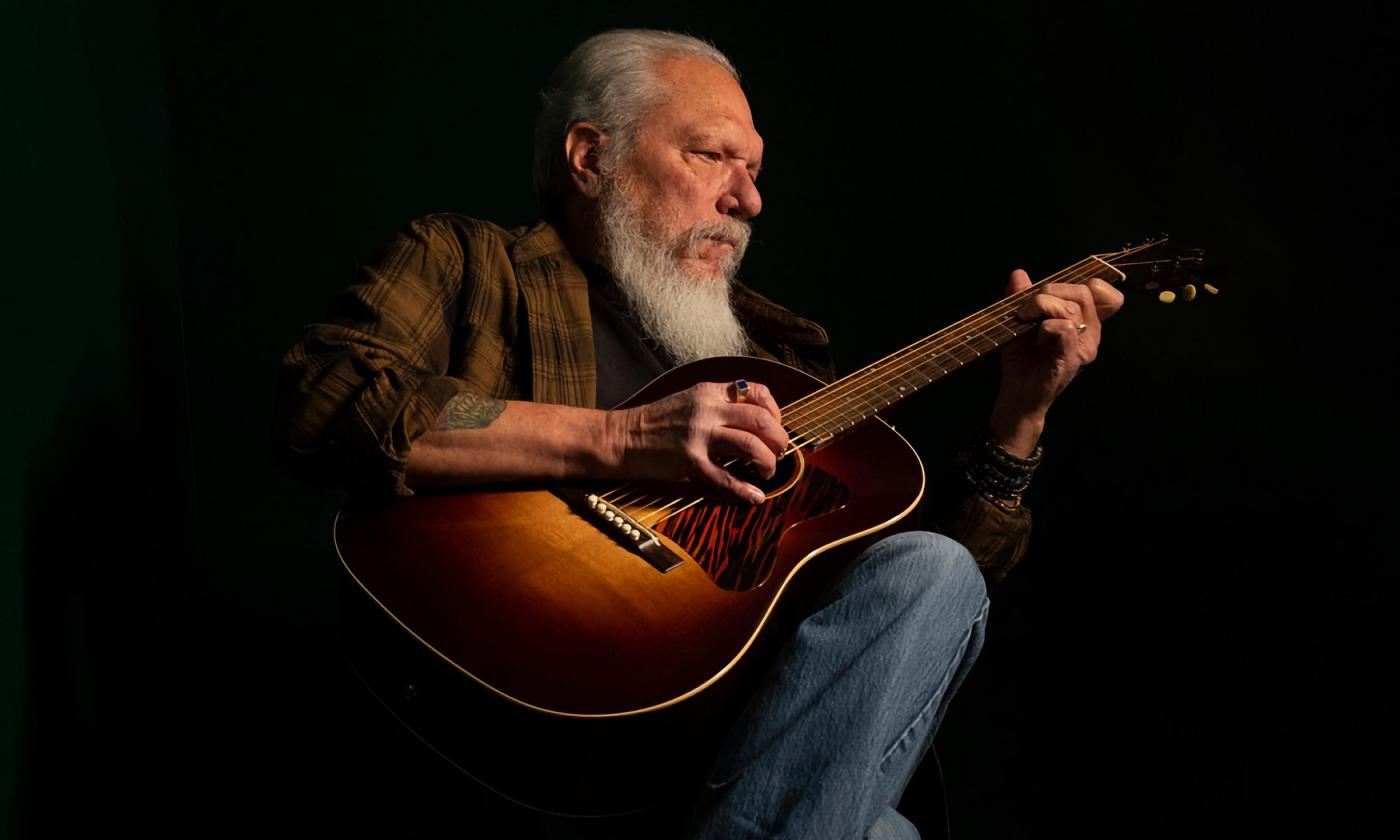 Jorman Kaukonen in jeans and a plaid shirt, strumming an acoustic guitar in a darkened stage.