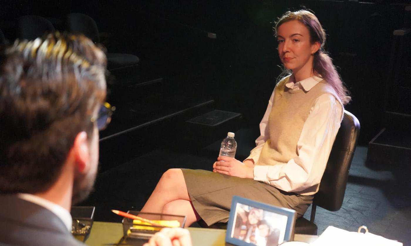 Woman sits, facing a man at a desk on a darkened stage