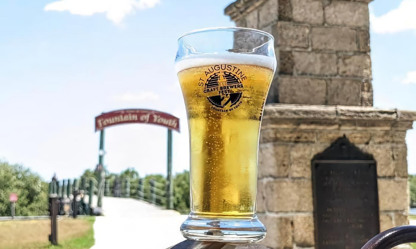 A glass of beer with the St. Augustine Craft Brewery Festival logo, in front of the Fountain of Youth