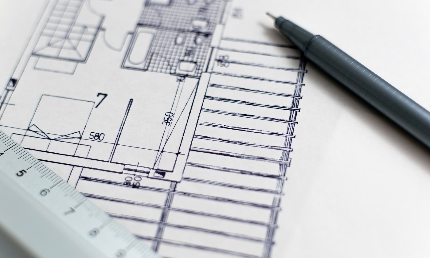 A close up full color image of a drafted drawing of a home's design. A pen and ruler are laid over the paper for aesthetic appeal.