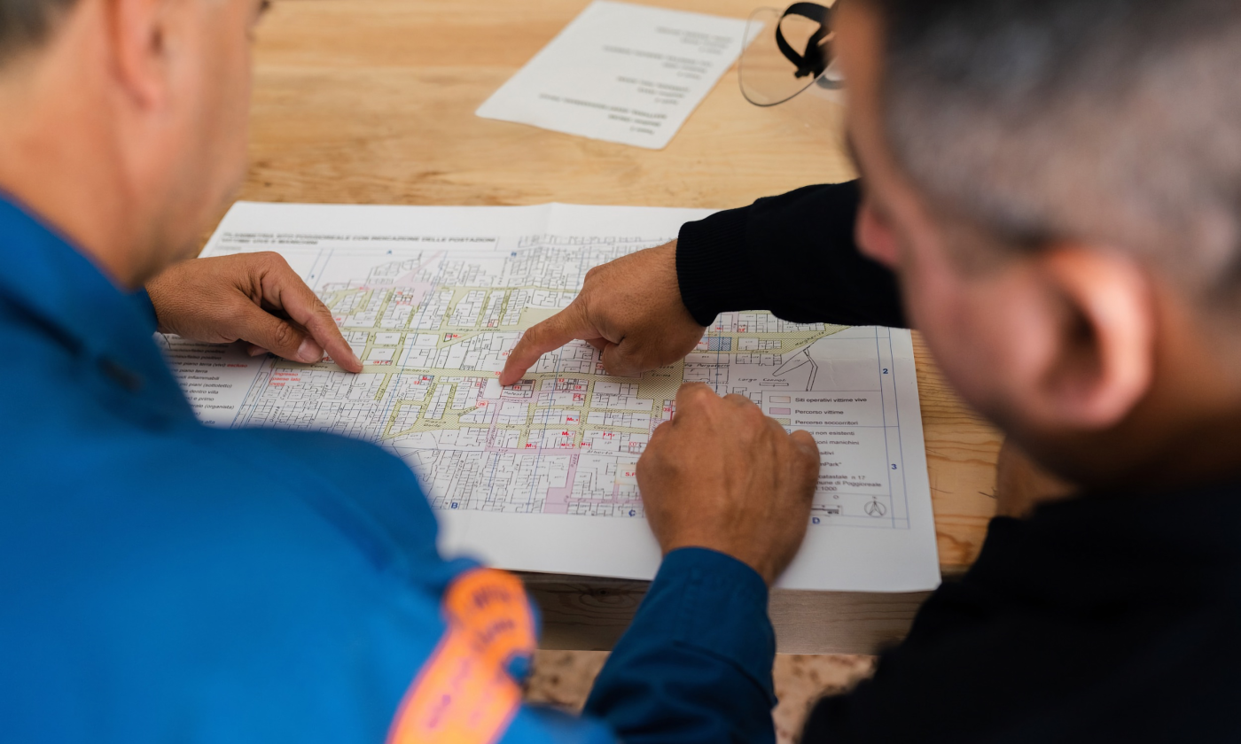 A full color image of two men facing a desk full of construction planning documents. They point to the paper, assumedly making a plan. 