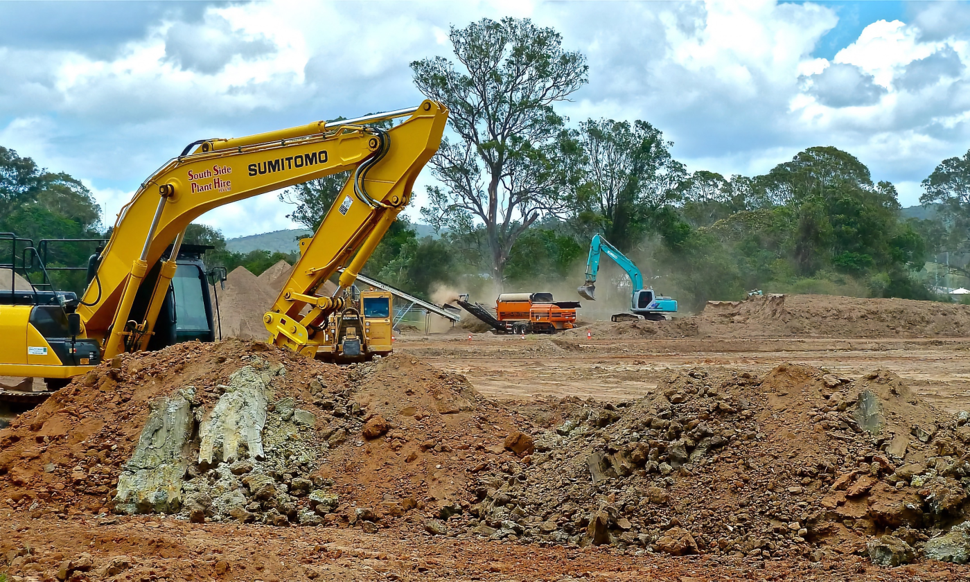 A full color image of a yellow bulldozer in a cleared landscape surrounded in piles of dirt. There are trees and a few other colorful machines in the background.