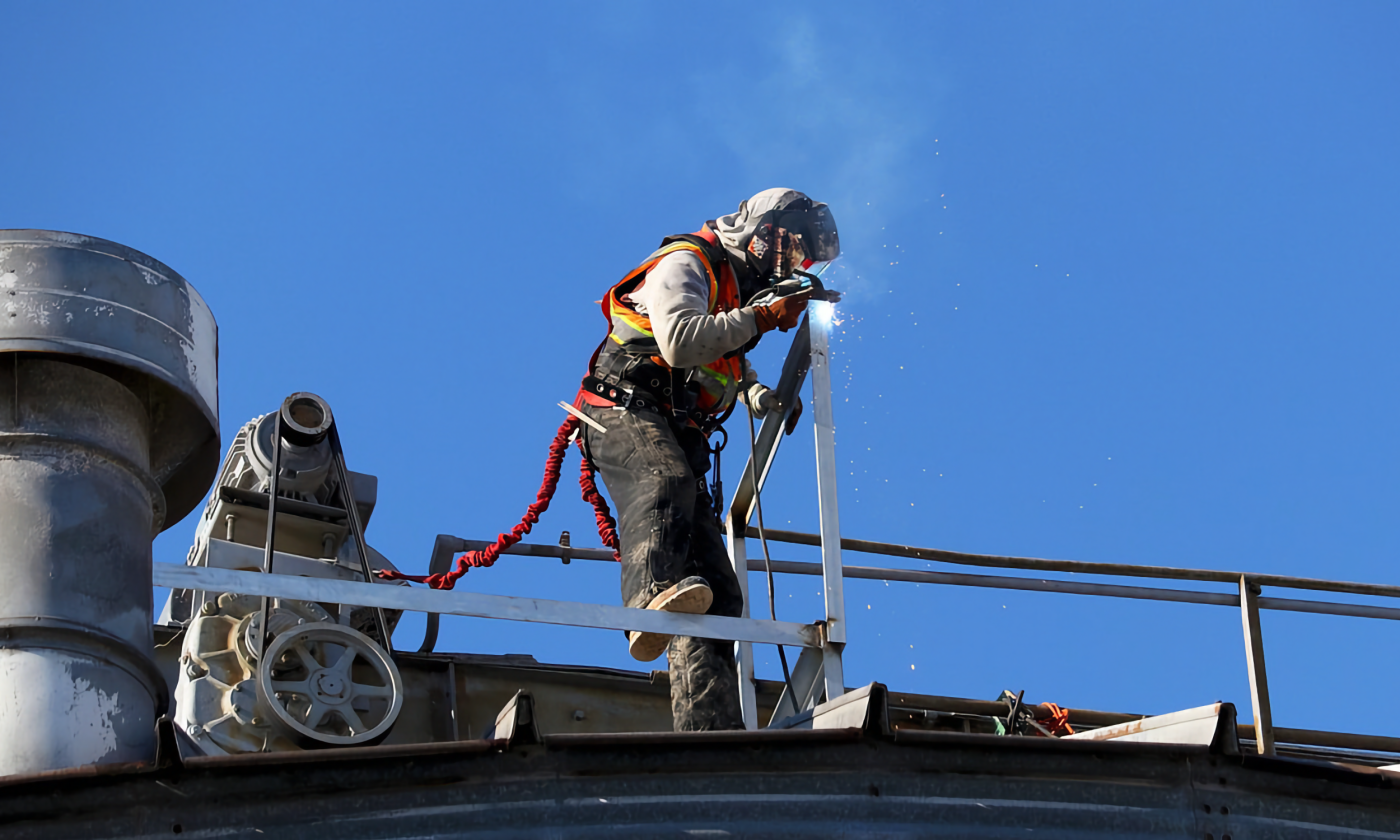 A full color picture of a construction worker on a metal roof above the photographer. He is dressed fully in protective gear and is hooked to the roof with a safety chord as he welds. Sparks and smoke rise from his tools into the blue sky behind him.