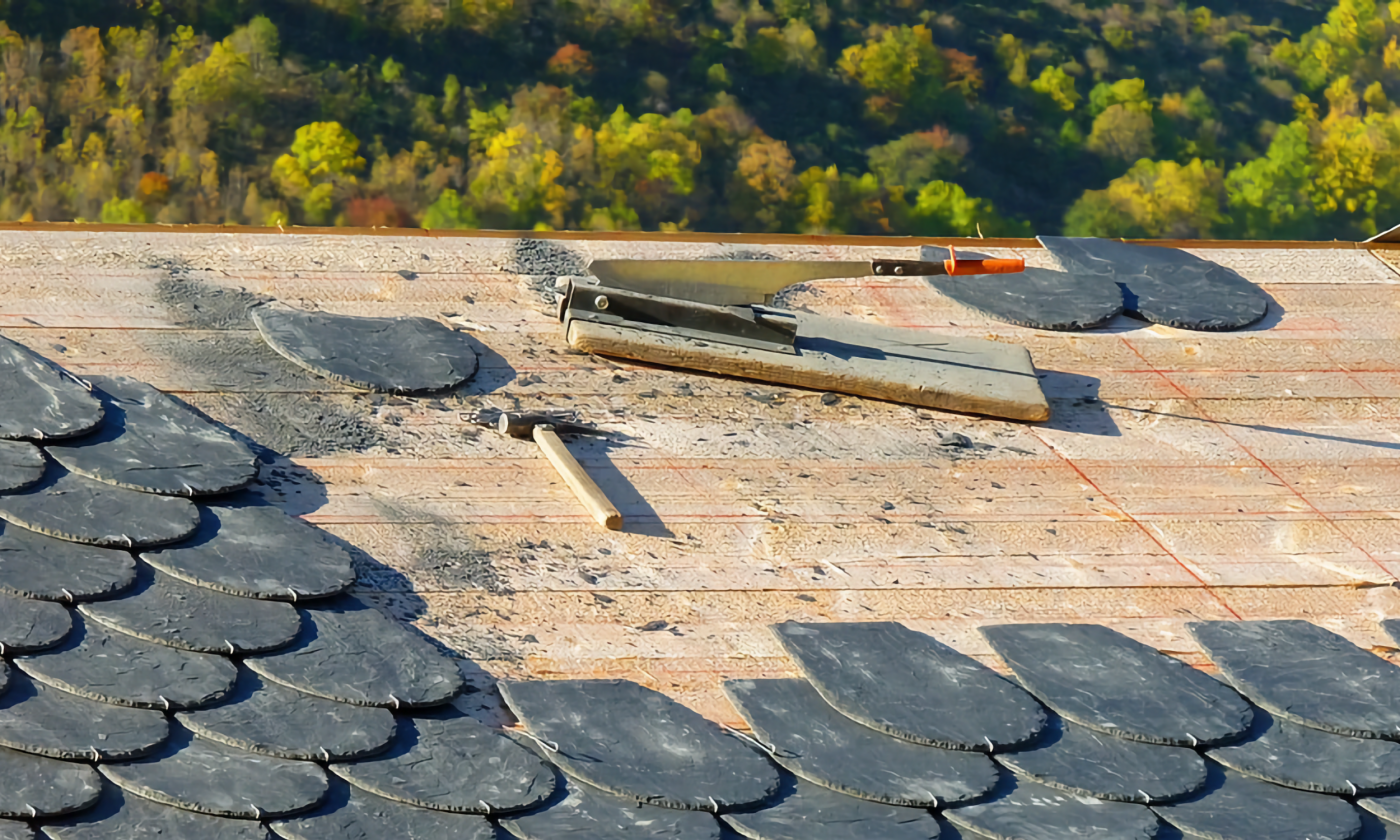 A full color image of a roofer's tools (small wooden hammer and tiles and scraper) on a semi-tiled roof. The shingles (tiles) are ovular and gray. Trees can be seen past the roof ridge in the background.