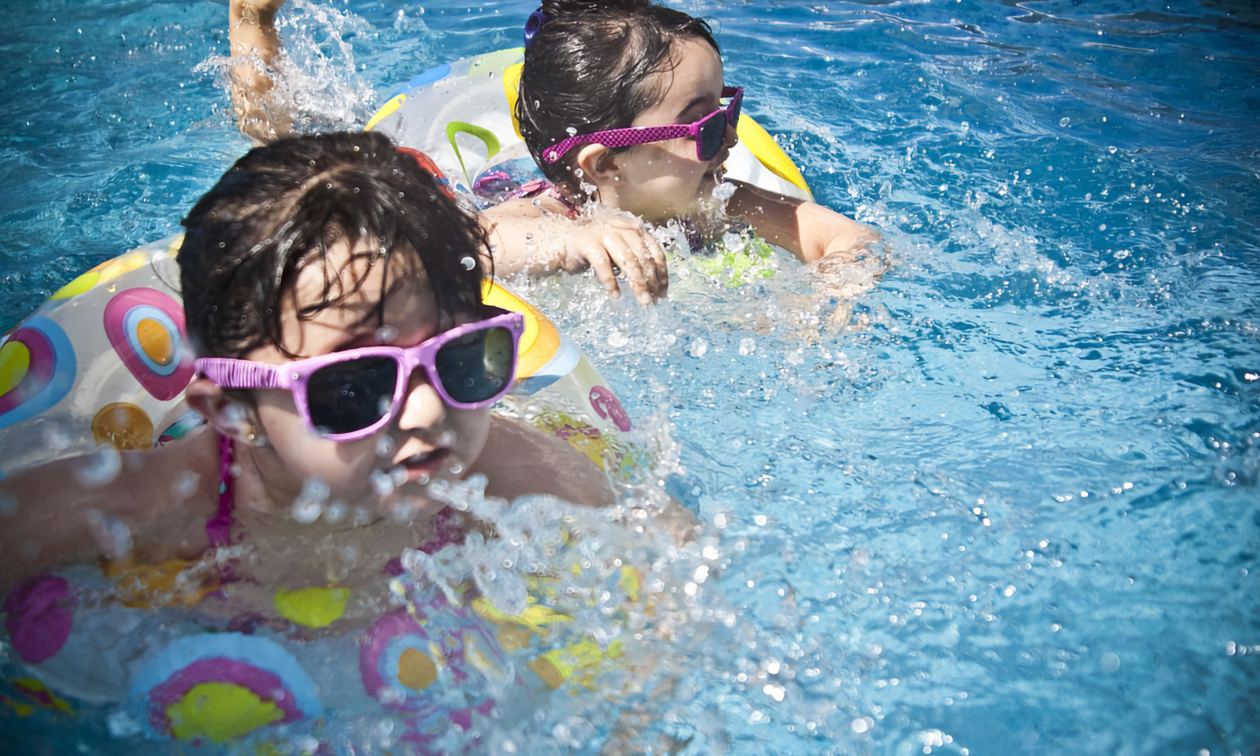 A full color image of two young dark haired girls in a swimming pool. They both use pink and yellow floaties.