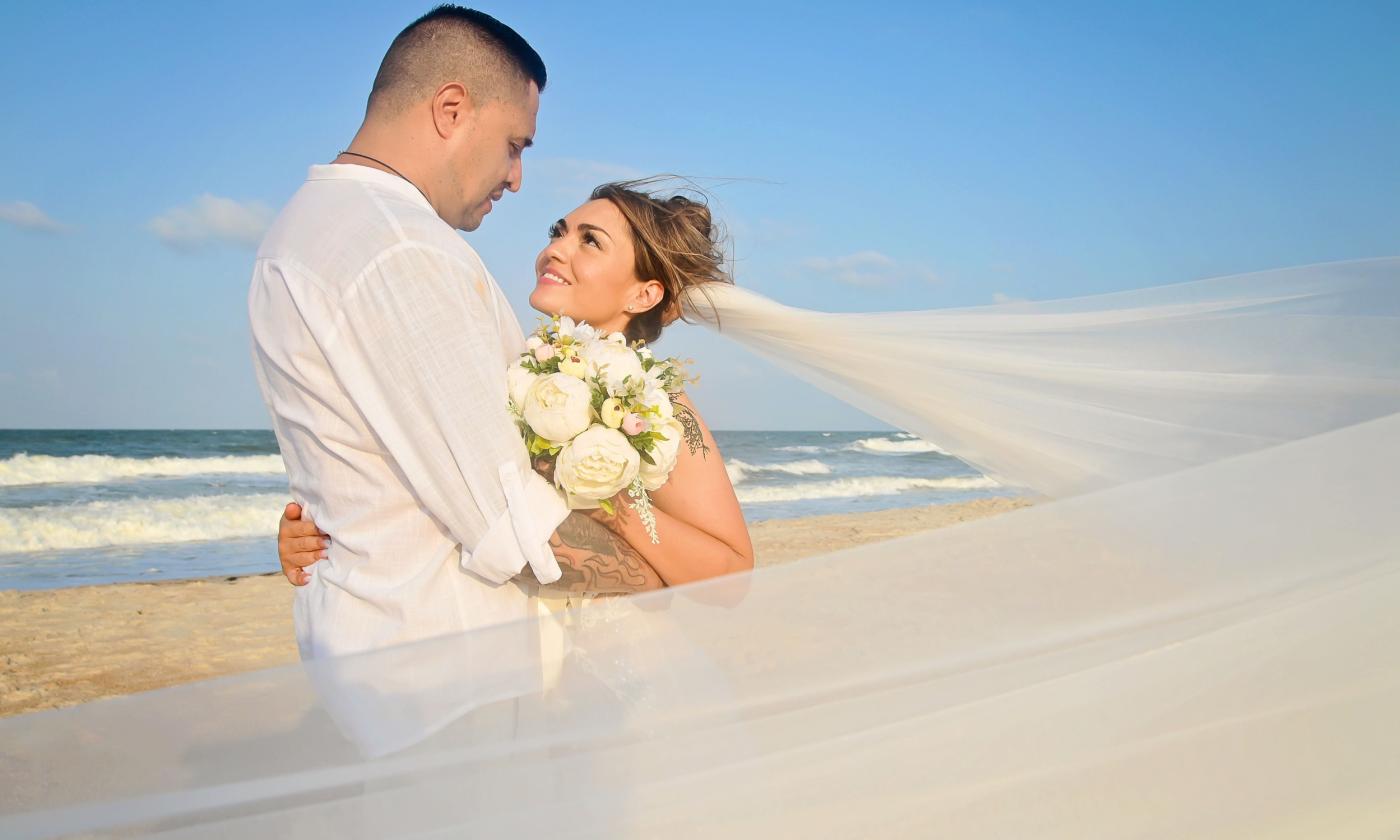 A bridal couple standing on the beach, surrounded by her wind-blown veil