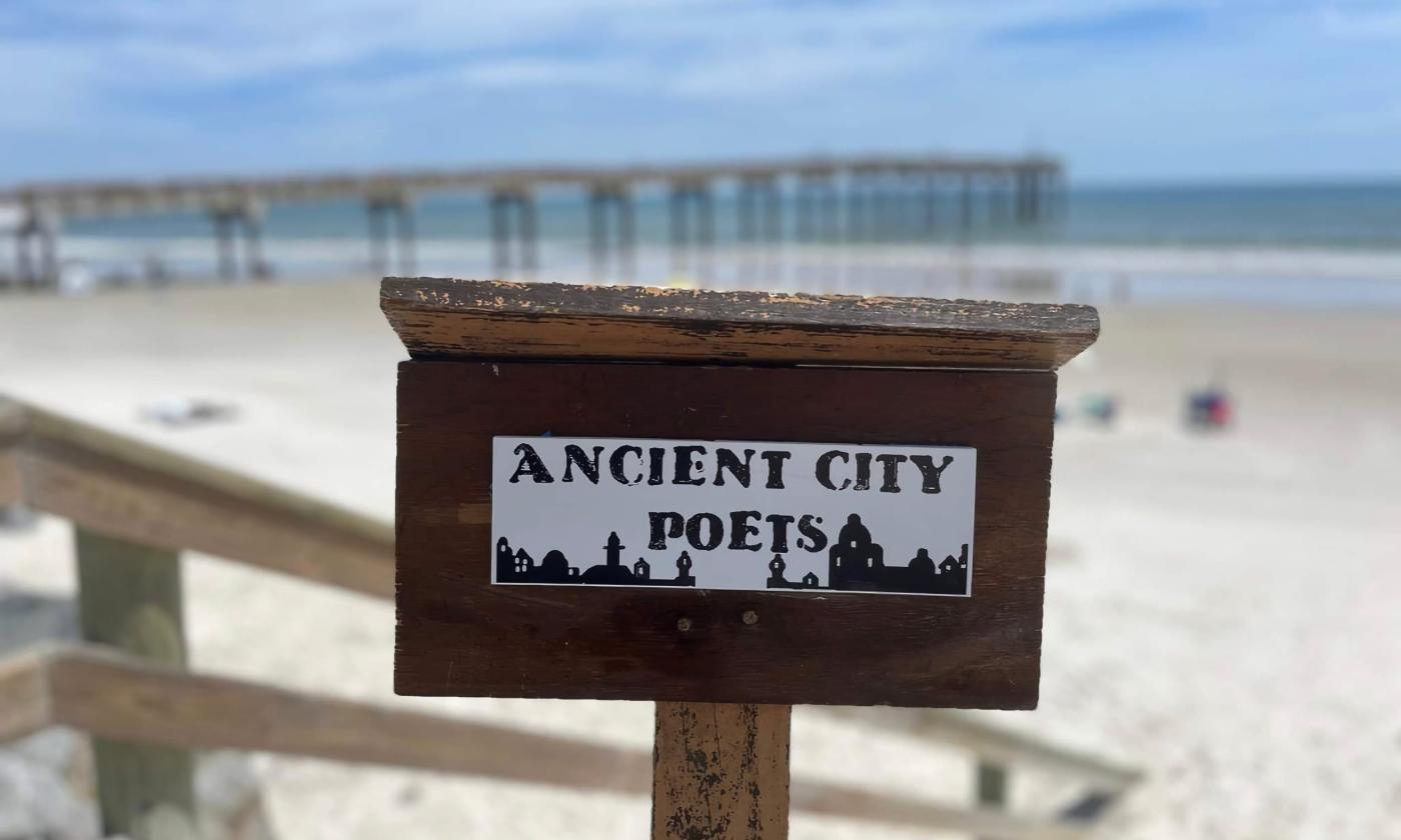 A podium with the Ancient City Poets logo (b&w) stands on a boardwalk with the St. Augustine Beach Pier and ocean in the background