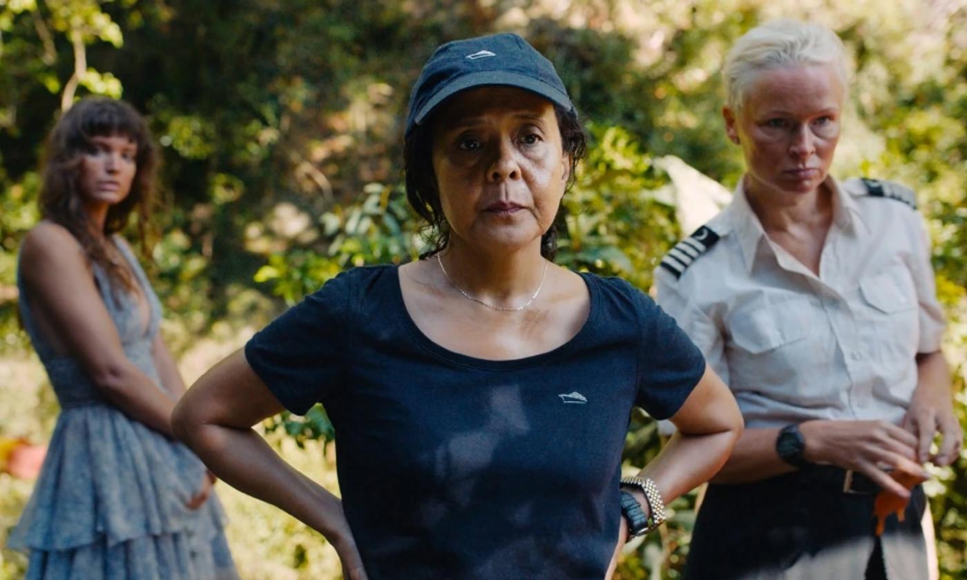 Actresses in the film Triangle of Sadness stare past the camera in front of a dense forest