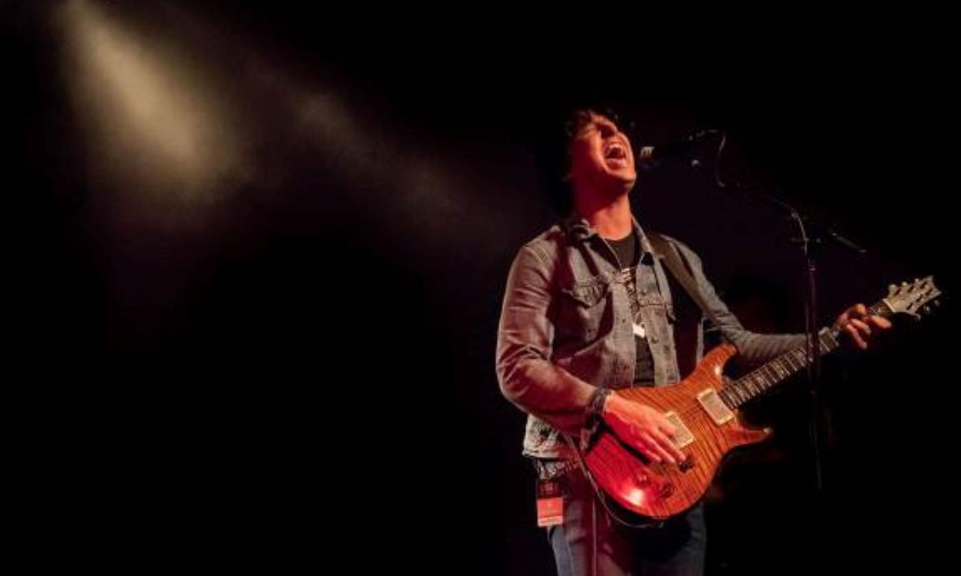 Davy Knowles holds his guitar and belts out lyrics on a dark stage with lights overhead