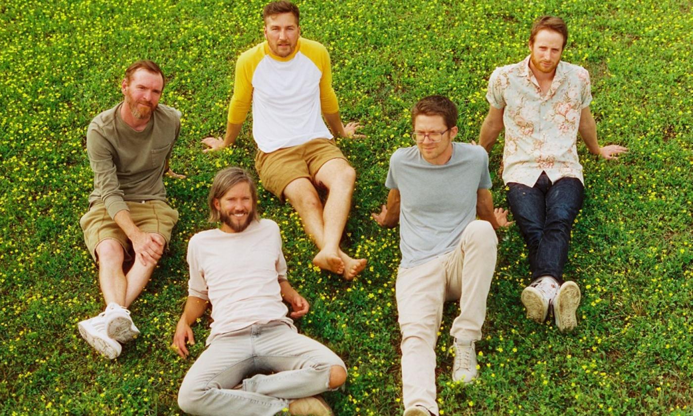 Members of the band Moon Taxi wear neutral colors and pose on lush green grass