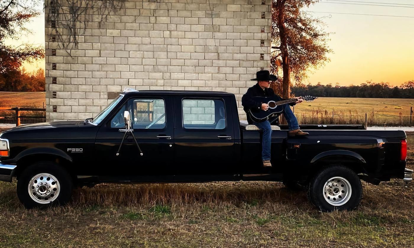 Clint Anglin, singer and songwriter, sitting with his guitar in the back of a black pick-up truck, near a field at sunset