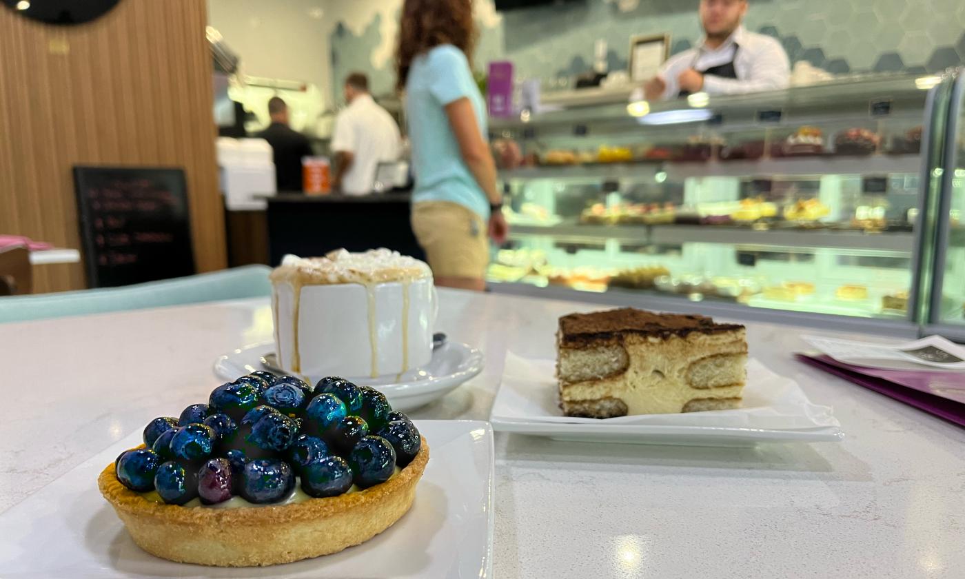 A blueberry pastry, tiramisu, and cappuccino in the foreground. Behind, the pastry counter at Cravings and More Bakery where customers browse delicious treats