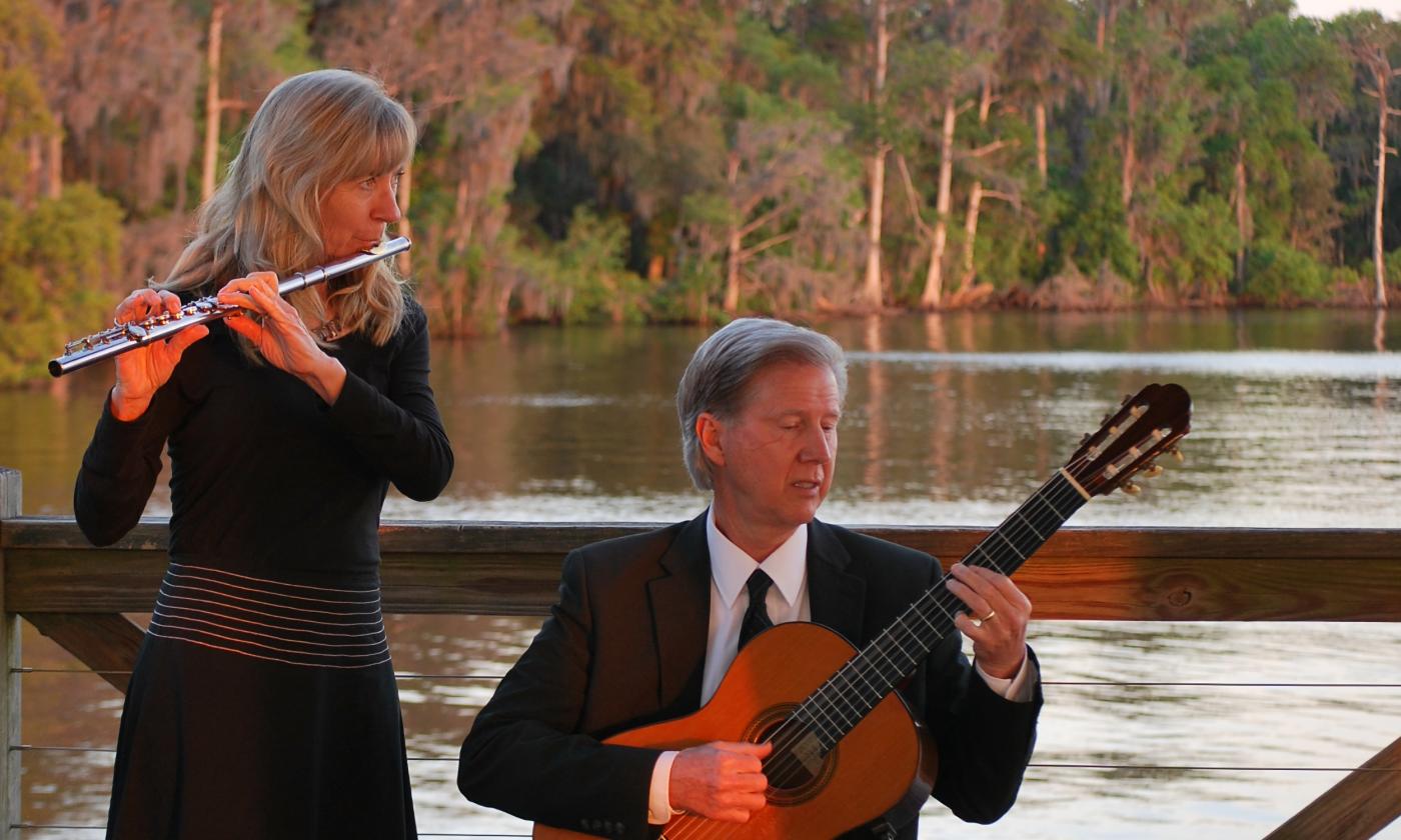 Laura and Chuck Rogers, playing their instruments at an outdoor setting on a river. Laura is standing and playing the flute and Chuck is seated playing the guitar