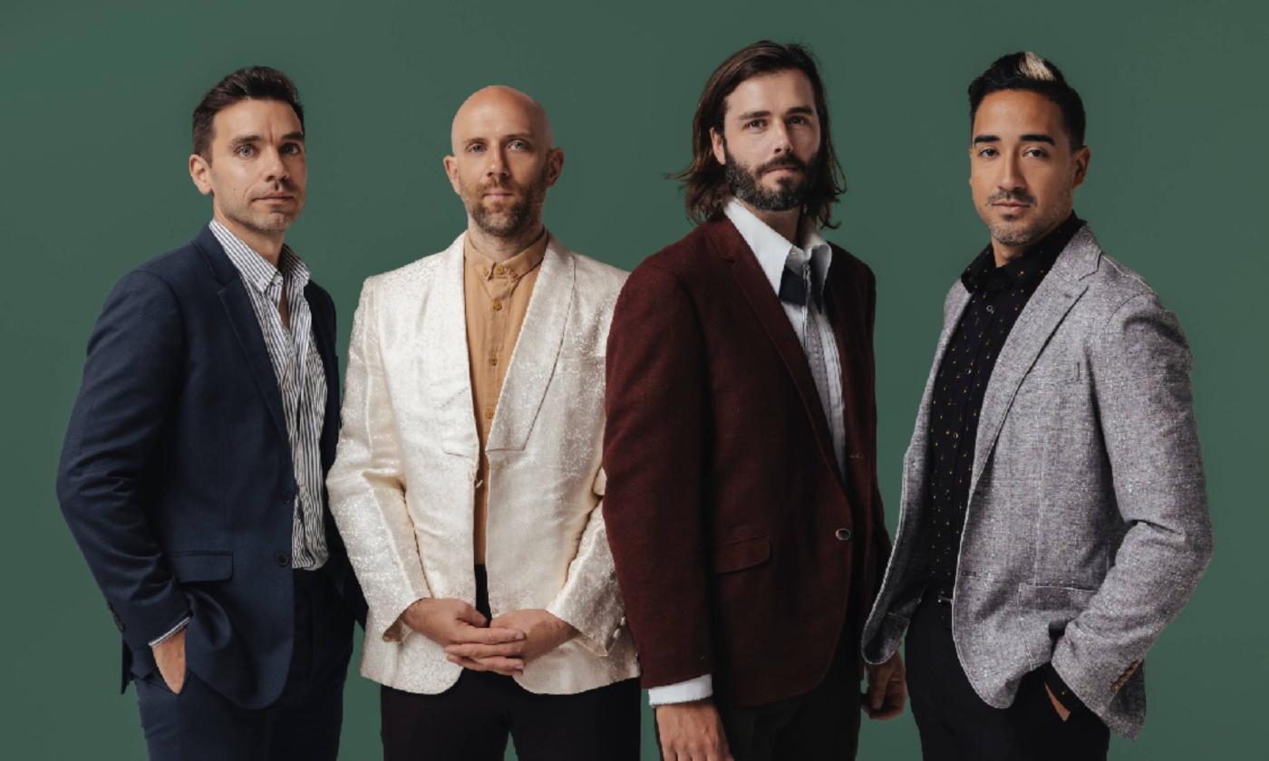 Four men (members of the band Lord Huron) stand shoulder to should looking at the camera against a green background. They are dressed in trendy suits